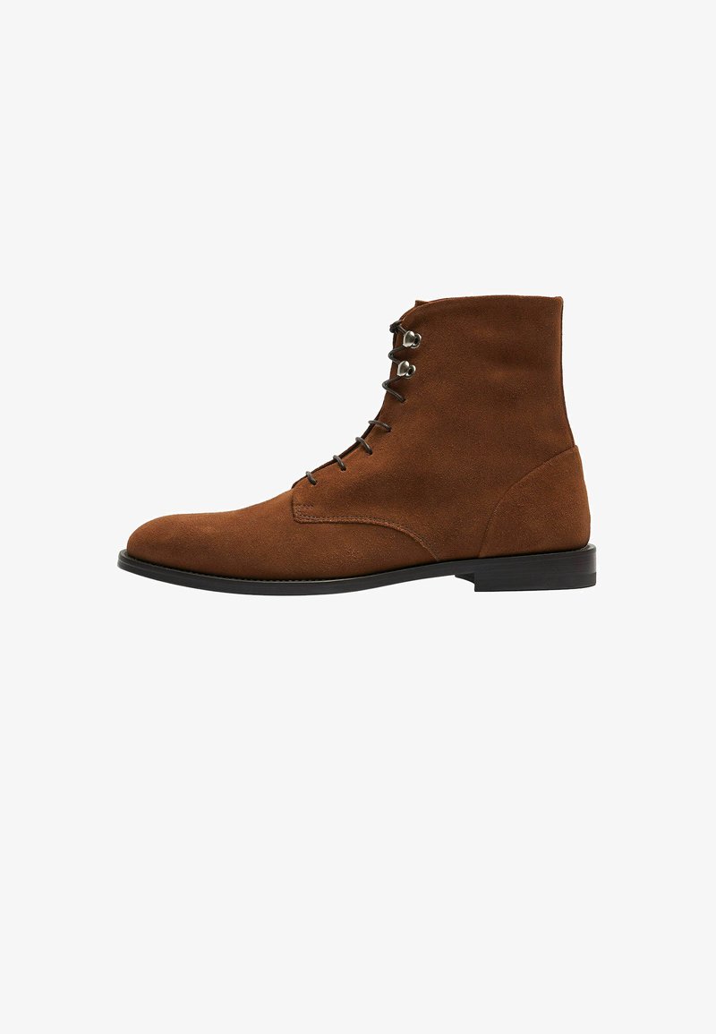 Bota de tobillo de ante marrón con punta redondeada y un bajo tacón negro. Presenta un diseño de cordones con seis ojales y una textura suave.