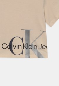 Camiseta beige con grandes letras "CK" superpuestas y el texto "Calvin Klein Jeans" impreso en la parte inferior frontal.