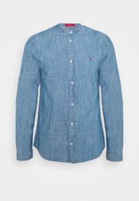 Světle modrá džínová košile s dlouhými rukávy, kulatým límečkem, předními knoflíky a jemným logem na hrudi. Měkká textura, neformální design.