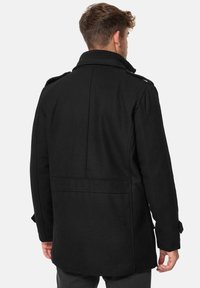 Abrigo negro hecho de tela con mezcla de lana; presenta un cuello alto, hombreras abotonadas y una parte trasera estructurada con una textura suave.