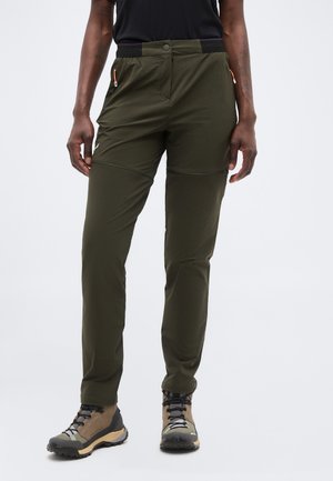 Személy olívazöld túranadrágot és bézs túracipőt visel, fehér háttéren áll.