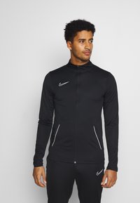 Zwarte sportjack met hoge kraag, lange mouwen, ritssluiting en wit Nike-logo op de borst. Heeft twee zijzakken met rits.