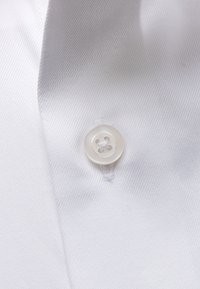 Valkoinen paitapusero, jossa on yksityiskohtana pyöreä neljäreikäinen nappi, ommeltu valkoisella langalla, korostaen kankaan rakennetta.