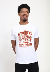 Bílý tričko s červeným grafickým designem, na kterém je kreslená postavička basketbalisty, slova "STREETS PRO GAME" a hravé basketbalové motivy.