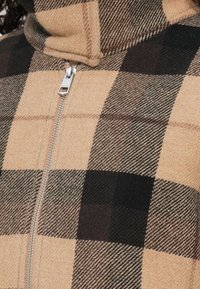 Sandfarbenes und dunkelbraunes Karo-Muster Reißverschluss-Jacke mit hohem Kragen und glatter Textur, ausgestattet mit einem sichtbaren Metallreißverschluss in der Mitte.