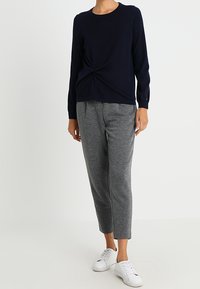 Haut à manches longues bleu marine avec un détail torsadé à l'avant, associé à un pantalon gris fuselé. Le modèle porte des baskets blanches. Design simple et décontracté.