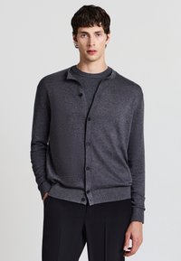 Cardigan grigio con colletto rotondo, chiusura a bottoni e polsini a coste. Realizzato in un tessuto a maglia testurizzato, indossato sopra una maglietta scura a girocollo.