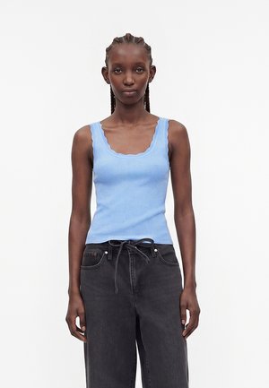 Vrouw met gevlochten haar draagt een lichtblauw geribd tanktopje en losse zwarte broek, staand tegen een effen witte achtergrond.