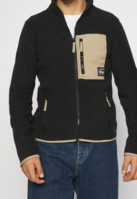 Czarna polarowa kurtka z beżową kieszenią na piersi, z zapięciem na zamek i dwoma bocznymi kieszeniami. Kontrastowe przeszycia i elastyczne mankiety dopełniają całość.