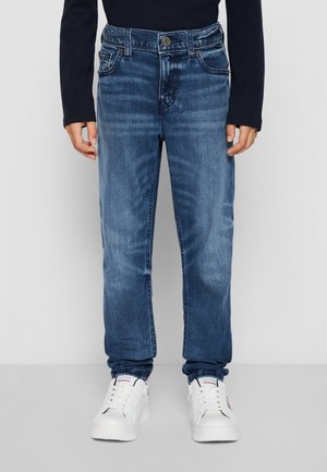 Niebieskie dżinsy z prostym krojem, widocznym przeszyciem, pięciokieszeniowym designem i zapięciem na guzik. Noszone z białymi trampkami.