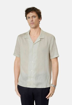 Man draagt een crème-kleurig shirt met korte mouwen en dunne verticale strepen, en zwarte broek, staat met ontspannen armen.