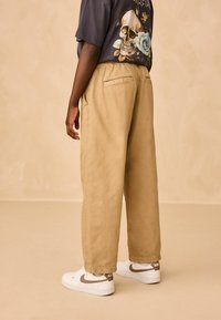 Pantaloni din bumbac bej, cu o croială lejeră, având un șnur la talie și buzunare laterale, asortați cu adidași albi.