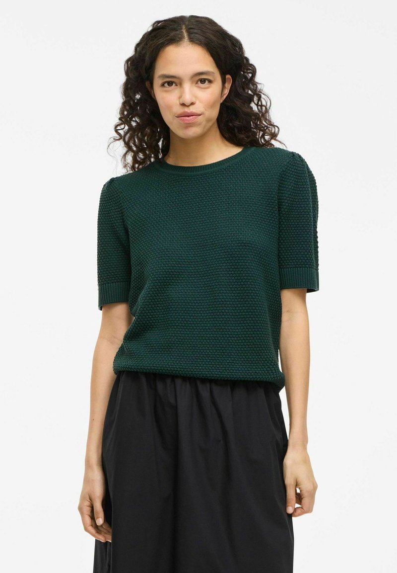 Top de punto de color verde oscuro y manga corta con patrón texturizado, cuello redondo y corte holgado, combinado con una falda negra.