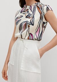 Blusa de manga corta con estampado floral en crema, rosa y marrón, con un escote fruncido y tejido suave, combinada con una falda blanca de lino.