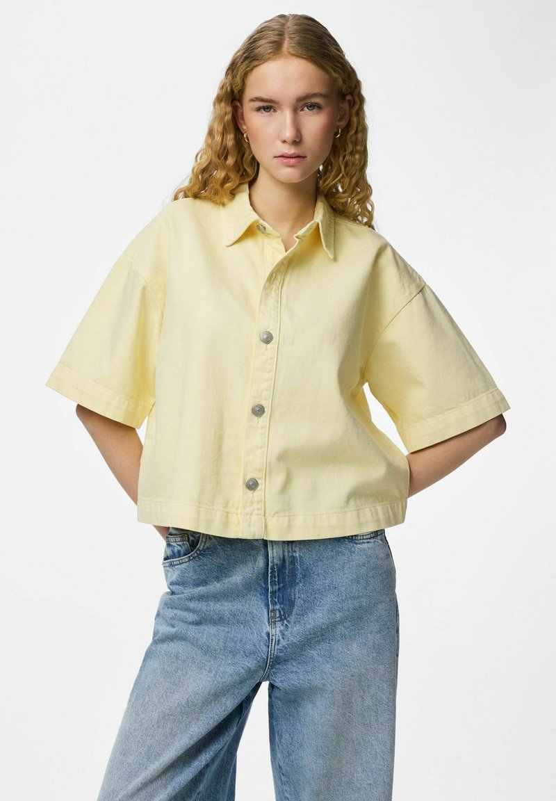Jeune femme aux cheveux bouclés portant une chemise ample à manches courtes boutonnée de couleur jaune pâle et un jean taille haute bleu clair.