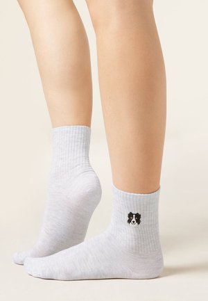Chaussettes blanches mi-mollet portées aux pieds, chaque chaussette présentant un petit emblème de visage de chien noir et blanc sur le côté extérieur au-dessus de la cheville.