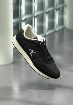 Czarne niskie sneakersy Calvin Klein z białą podeszwą i logo po boku, prezentowane na rozmytym tle nowoczesnego wnętrza.