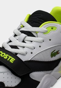 Baskets blanches et noires avec surfaces texturées, accents vert néon, lacets en tissu, et une bride avec le logo Lacoste.
