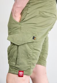 Pantaloni scurți cargo de culoare verde măsliniu, dintr-un material ușor, cu buzunare laterale, un buzunar la spate și o etichetă de marcă roșie și neagră pe tiv.