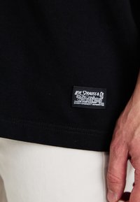 Svart bomull t-skjorte med et hvitt merke som har teksten "Levi Strauss & Co" og grafikk, synlige sømmer langs kanten, nærbilde av en hånd som holder den.