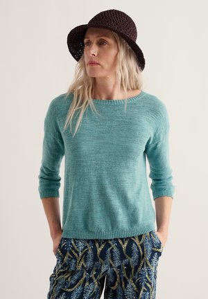 Frau trägt einen türkisfarbenen Strickpullover, schwarzen gewebten Hut und gemusterte blaue und grüne Hose, steht mit den Händen in den Taschen und schaut nach links.