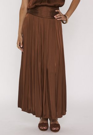 Femme portant une longue jupe plissée marron avec des sandales tressées assorties et des bagues et bracelets en or aux mains.