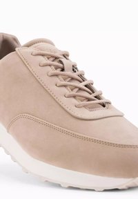 Beige mockasinsneaker med snörning, rund tå och vit gummisula. Har sömdetaljer och en slät yta.