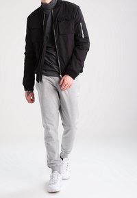 Black bomber jacket with two front pockets and a zippered arm pocket, paired with light gray sweatpants and white sneakers.