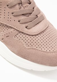 Jasnobrązowy zamszowy sneakers z perforowaną cholewką, okrągłym noskiem i dopasowanymi sznurowadłami. Posiada białą gumową podeszwę i teksturowane detale.