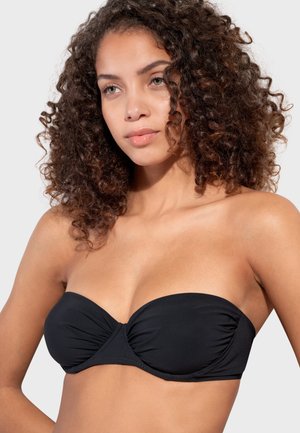 Donna con capelli ricci che indossa un top bikini bandeau nero senza spalline su uno sfondo chiaro e semplice.