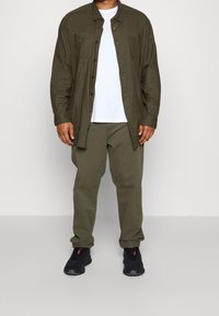 Camicia verde a maniche lunghe sopra una t-shirt bianca, abbinate a pantaloni verdi e sneakers nere. La camicia presenta taschini sul petto e una vestibilità comoda.