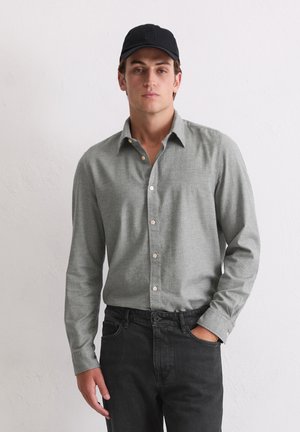 Graues Hemd mit Knopfverschluss, strukturiertem Muster, umgeschlagenem Kragen und cremefarbenen Knöpfen, kombiniert mit schwarzen Jeans und einer schwarzen Mütze.
