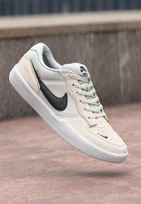 Nike SB tenisice u svijetlo sivoj i kremasto-bijeloj boji, s gornjim dijelom od antilopa, crnim swoosh logotipom, teksturiranim detaljima i bijelim gumenom potplatom.