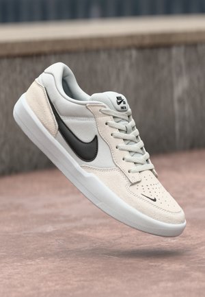 Sneaker Nike SB w jasnoszarym i kremowym kolorze, z zamszowym wierzchem, czarnym logo Swoosh, teksturowanymi detalami i białą gumową podeszwą.