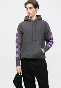 Hanorac gri cu buzunar frontal și șnur ajustabil. Prezintă modele grafice mov pe ambele mâneci. Material moale, cu textură. Pantaloni negri.