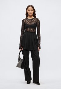 Μαύρο μακρυμάνικο τοπ από δαντέλα πάνω από μαύρο μπούστο, συνδυασμένο με πλισέ μαύρη φούστα και παντελόνι με φαρδιά πόδια. Τα αξεσουάρ περιλαμβάνουν μια μαύρη τσάντα χειρός.