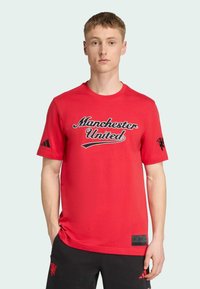 Camiseta de algodón roja con "Manchester United" en guion negro, con el logo negro de Adidas en la manga izquierda y una etiqueta en el costado.