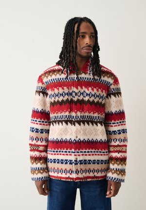 Homme avec de longues dreadlocks portant une veste polaire à motifs rouge, blanc, bleu et marron, et un jean bleu, debout devant un fond uni.