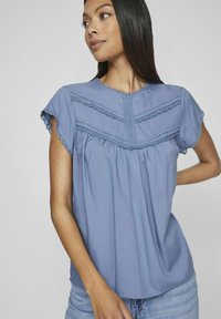 Blusa azzurra a maniche corte con dettagli in pizzo, arricciature e scollatura rotonda. Tessuto leggero che offre una texture morbida.