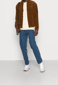 Giacca di velluto a coste marrone, maglione crema, jeans blu, sneakers bianche con suola in gomma. Borsa appesa a una spalla, in piedi su uno sfondo grigio.