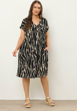 Frau trägt ein schwarz-beiges, zebramusterndes V-Ausschnitt-Kleid mit kurzen Ärmeln und goldenen Keilsandalen, steht vor einer schlichten Wand.