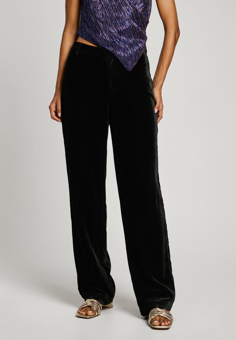 Femme portant un pantalon large en velours noir, un haut asymétrique violet brillant et des sandales dorées à brides ouvertes aux orteils, debout devant un fond uni.