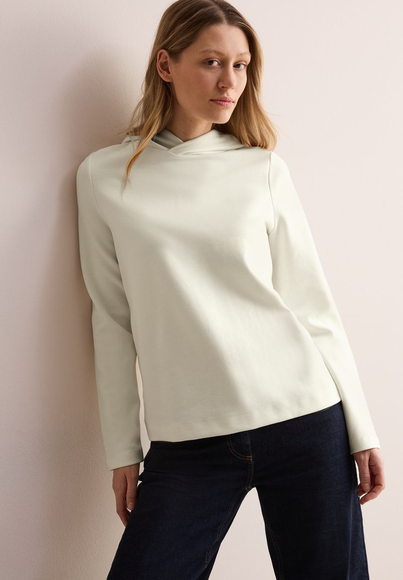 Blusa de manga larga verde claro con cuello estilo cowl, hecha de una tela suave, combinada con jeans azul oscuro con costuras amarillas.