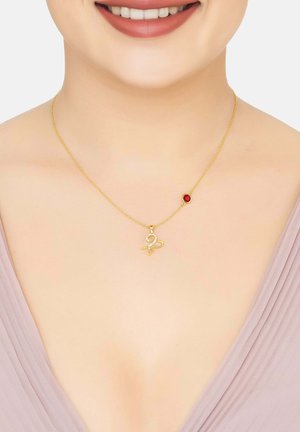 Gold necklace featuring a swirl pendant with a small red accent stone, displayed on a bare neckline against a soft, draped fabric.