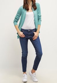 Cárdigan de canalé turquesa, top blanco sin mangas, vaqueros azul oscuro, zapatillas blancas. Ajuste slim con diseño de cárdigan abierto y sin patrones visibles.