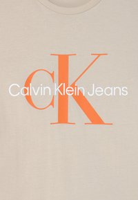 Beżowa bawełniana koszulka z dużą pomarańczową grafiką "CK" oraz białym napisem "Calvin Klein Jeans". Krótkie rękawy i okrągły dekolt.