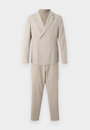 Giacca sartoriale beige a doppio petto con due tasche frontali, abbinata a pantaloni attillati coordinati. Linee pulite e design su misura.