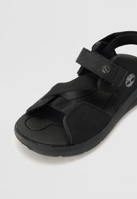Černé sandály s nastavitelnými pásky, texturovanou stélkou, polstrovanou podrážkou a detailem loga na boku. Lehký a odolný design.