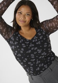 Haut noir à motifs floraux avec des manches longues transparentes, un corps texturé arborant un motif de fleurs subtil, et un décolleté en scoop. Associé à un pantalon gris.