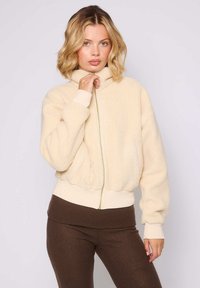 Giacca zip-up beige e pelosa con colletto alto, polsini e orlo a coste, con una cerniera verde chiaro. Indossata sopra leggings marroni.